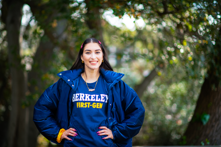 student smiling with her hands on her hips in front of green foilage; shirt reads Berkeley First Gen