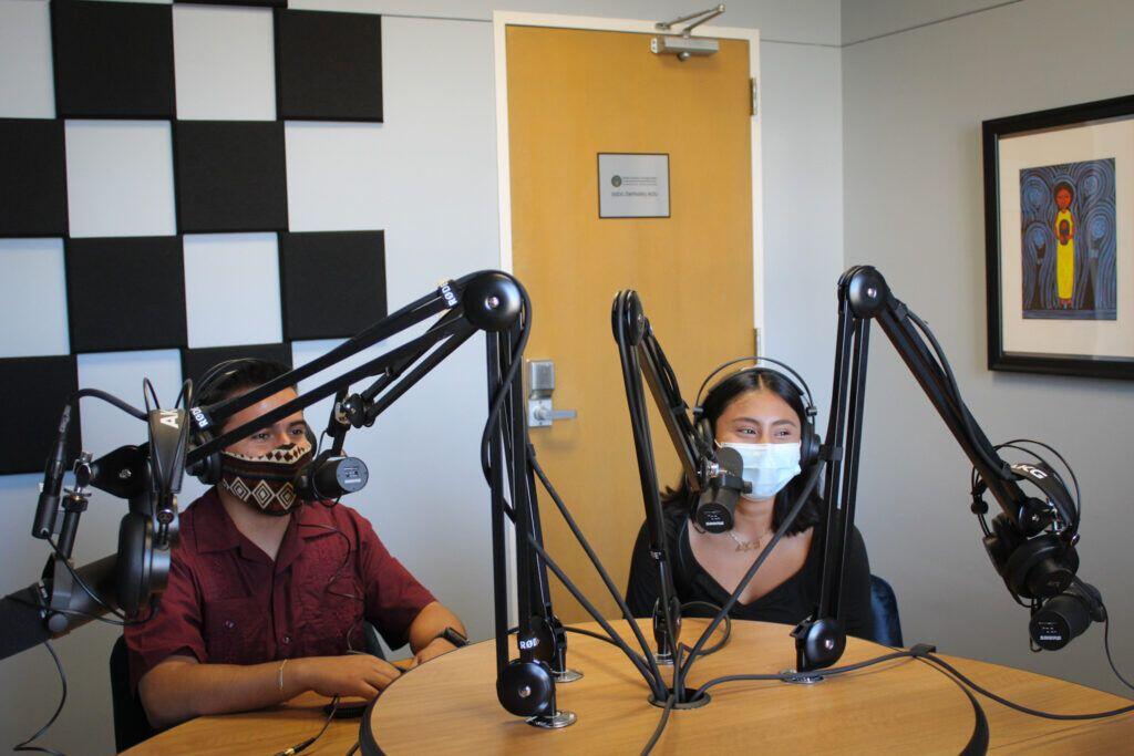 Two students wearing masks in front of microphones in a podcast studio