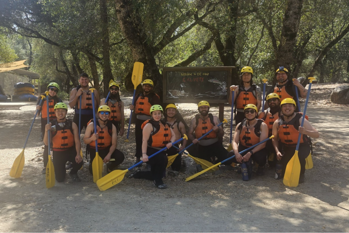 Veteran and military-connected students, staff, and alumni posing for a photo wearing personal protective equipment prior to getting on the South Fork of the American River to go whitewater rafting.