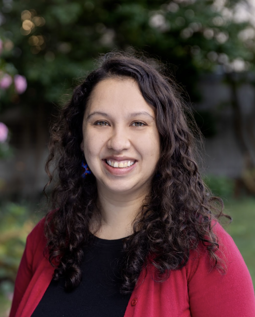A smiling woman with long, curly dark hair stands outdoors in front of a blurred background of green trees and pink flowers. She is wearing a black top with a red cardigan and blue beaded earrings.