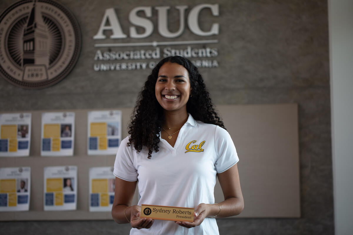 Sydney Roberts, the new ASUC president, holds a small sign with her name on it and her title. She is in Eshleman Hall standing in front of a wall that says ASUC with the ASUC symbol on it.