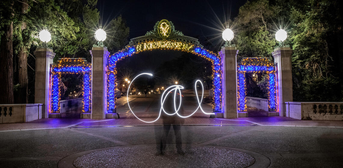 sather gate at night with yellow and blue lights with light art spelling cal in the center
