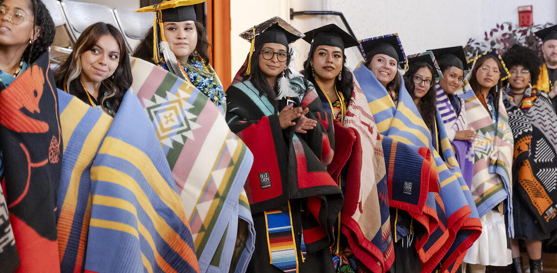 Graduating Native students stand in a line wearing their graduation caps and gowns and gorgeous Native blankets wrapped around t